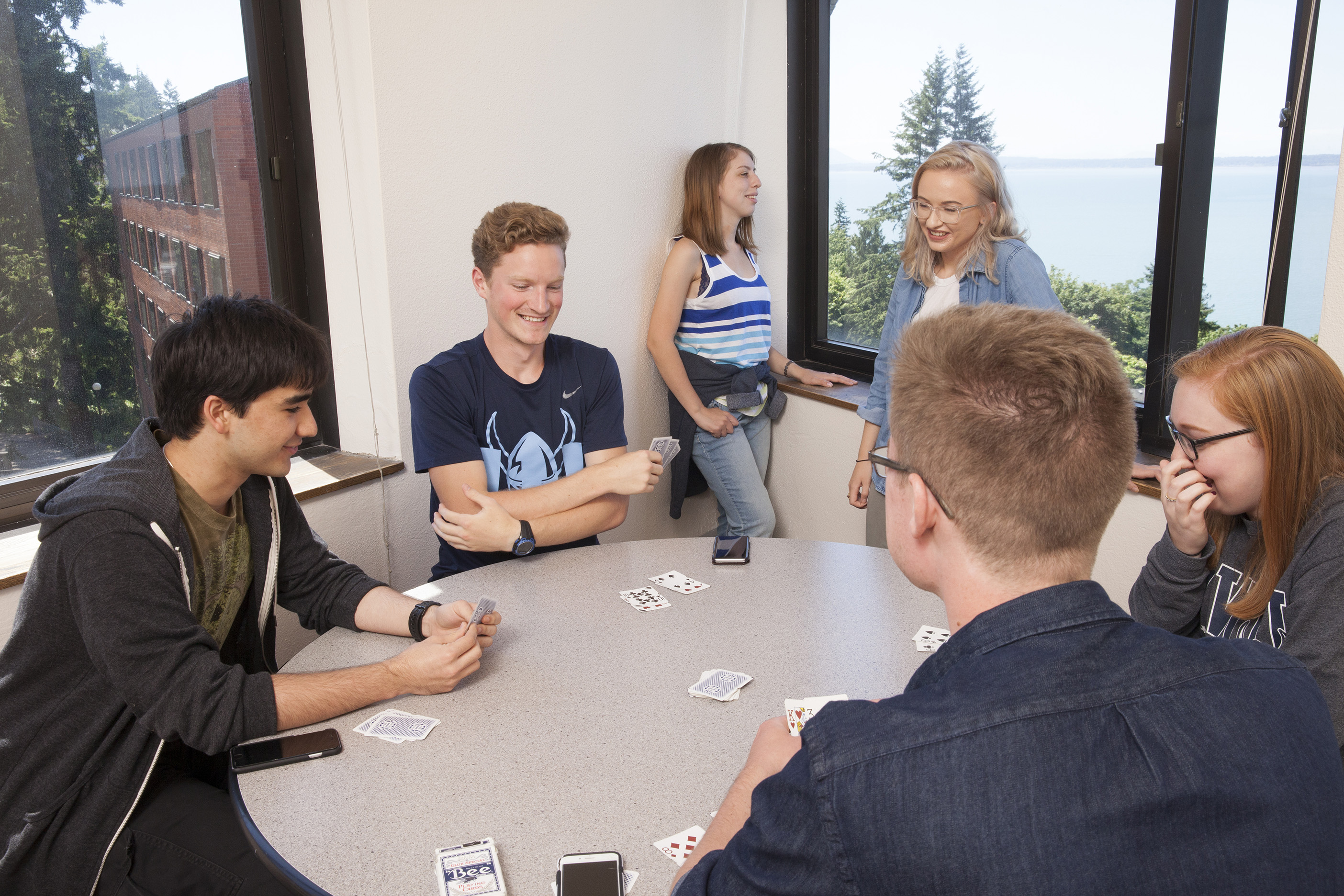 Four students play cards in the foreground while two other students sit on the window sill in the background. Taken in a Nash Hall Lounge.